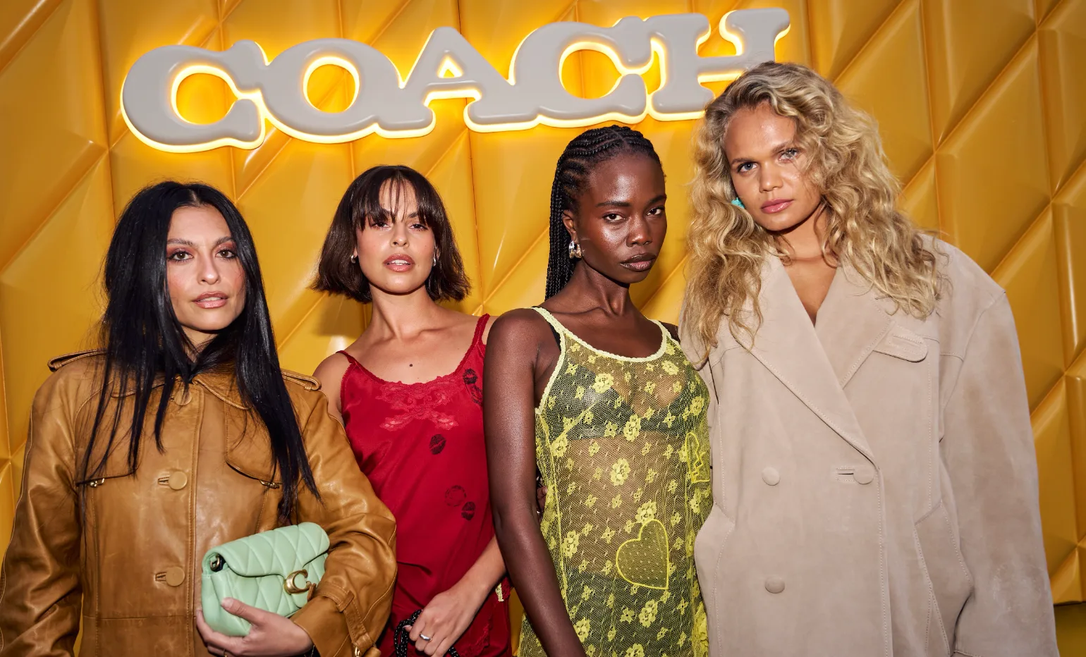 Four women posing in front of a yellow background with the illuminated Coach logo, wearing fashionable outfits.